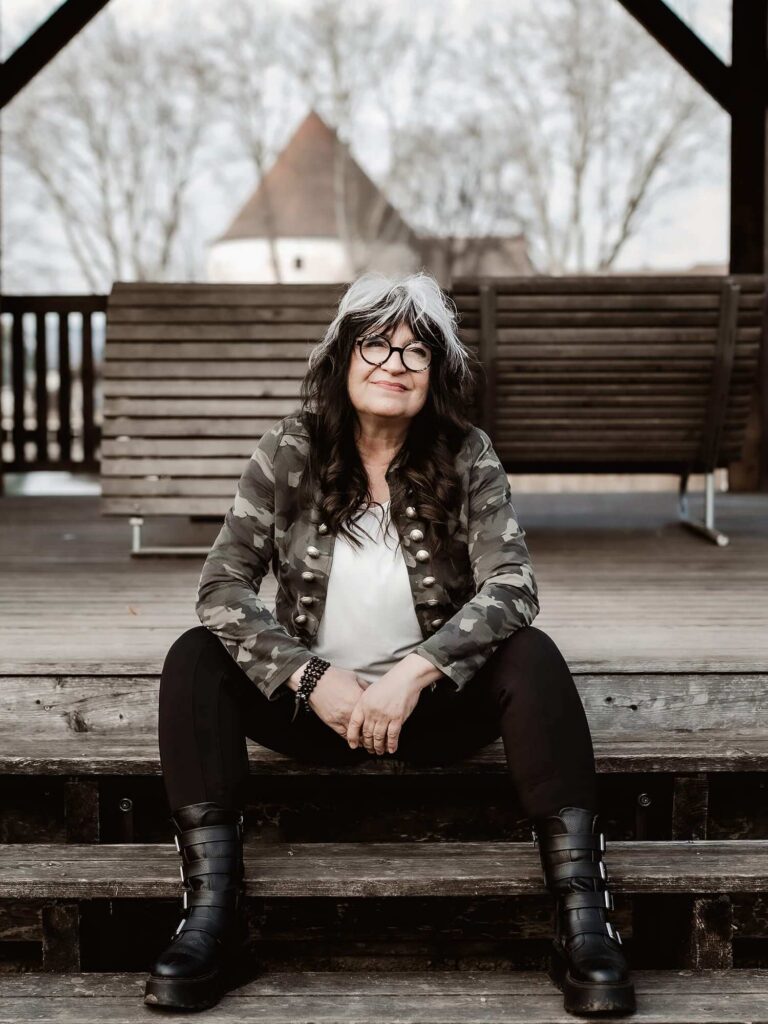 Dorothea „Doro“ Jäger sitzt entspannt auf Holzstufen, trägt Camouflagejacke und schwarze Stiefel. Authentisches Porträt der Gründerin des BAD-ASS Bootcamps für Lehrlinge.